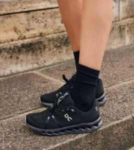 Feet in black On running sneakers with thick wavy soles, wearing black socks, standing on stone outdoor steps.