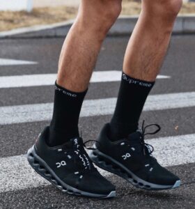 Runner wearing black On running shoes and black Supreme socks, standing on a street crosswalk with white lines.