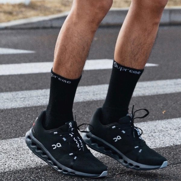 Runner wearing black On running shoes and black Supreme socks, standing on a street crosswalk with white lines.