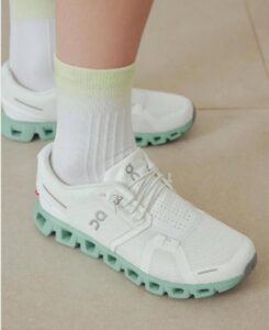 White running sneaker with a mint-green sole worn with white socks, shown from the side on a tiled floor.
