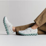 White On running shoes with a mint-green, chunky sole shown in a side view with brown pants nearby.