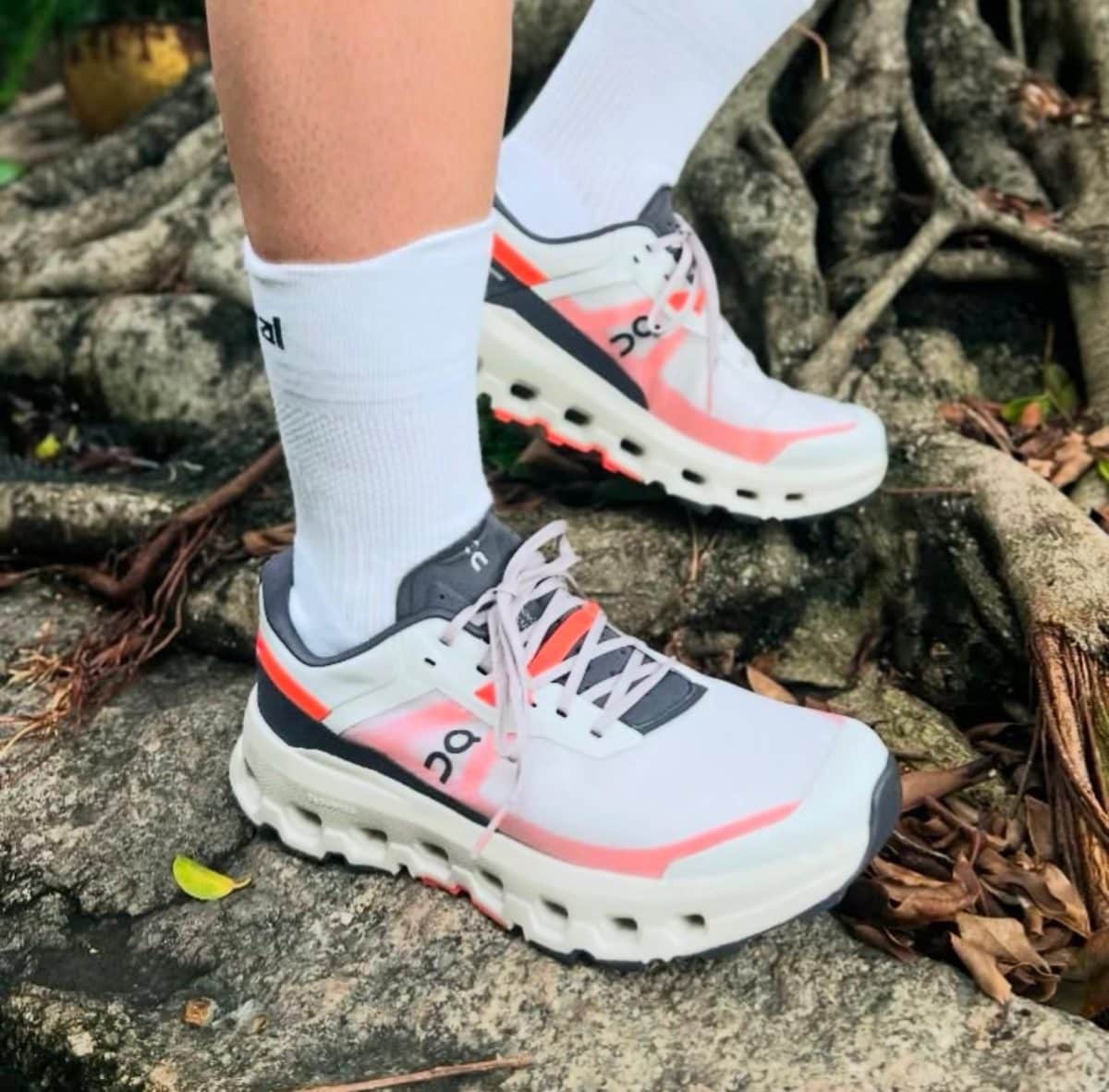 White and pink On running shoes with thick soles, worn with white socks, stepping on rocky outdoor terrain.