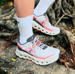 White and pink On running shoes with thick soles, worn with white socks, stepping on rocky outdoor terrain.