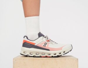 White and pink On running shoe on a wooden block, worn with a white crew sock.