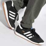 Black Adidas sneakers with white stripes and gum sole, being worn by a person wearing olive-green pants