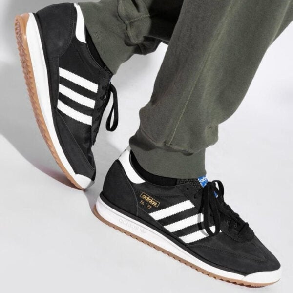 Black Adidas sneakers with white stripes and gum sole, being worn by a person wearing olive-green pants