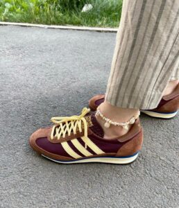 Person wearing maroon and tan sneakers with cream stripes and yellow laces, standing on pavement; ankle is adorned with a seashell beaded anklet; striped light-colored pants.