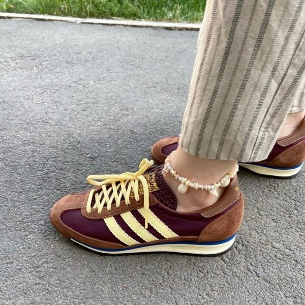 Person wearing maroon and tan sneakers with cream stripes and yellow laces, standing on pavement; ankle is adorned with a seashell beaded anklet; striped light-colored pants.
