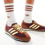 Close-up of feet wearing burgundy and tan Adidas sneakers with cream stripes, paired with white crew socks featuring red stripes, standing on a white surface.