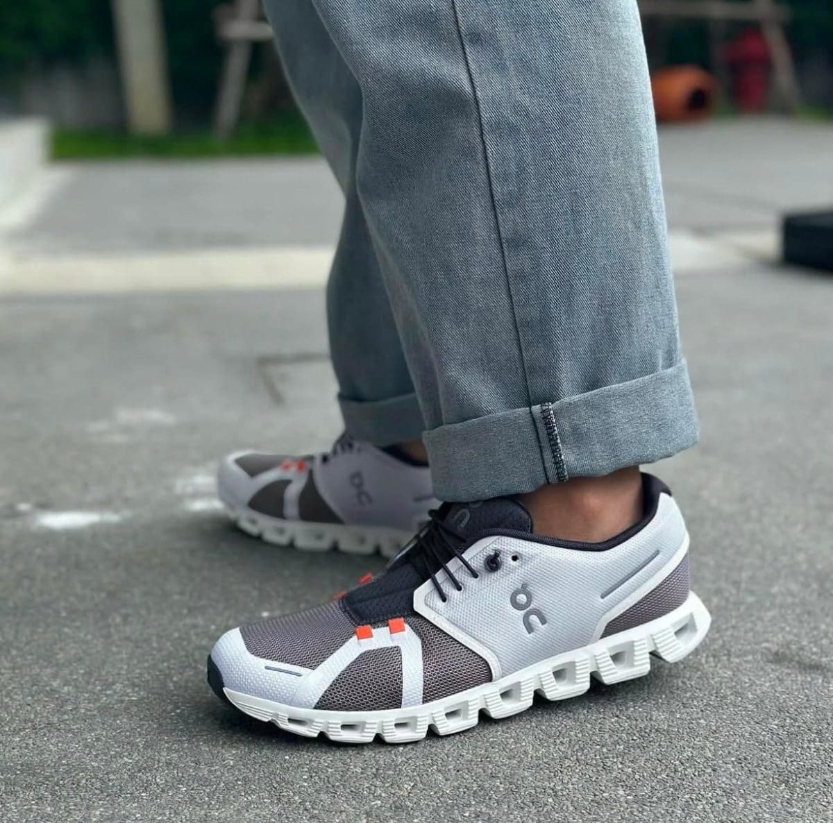 Gray and white running shoes with orange accents on a person’s feet, rolled-up jeans, standing on pavement in a park area