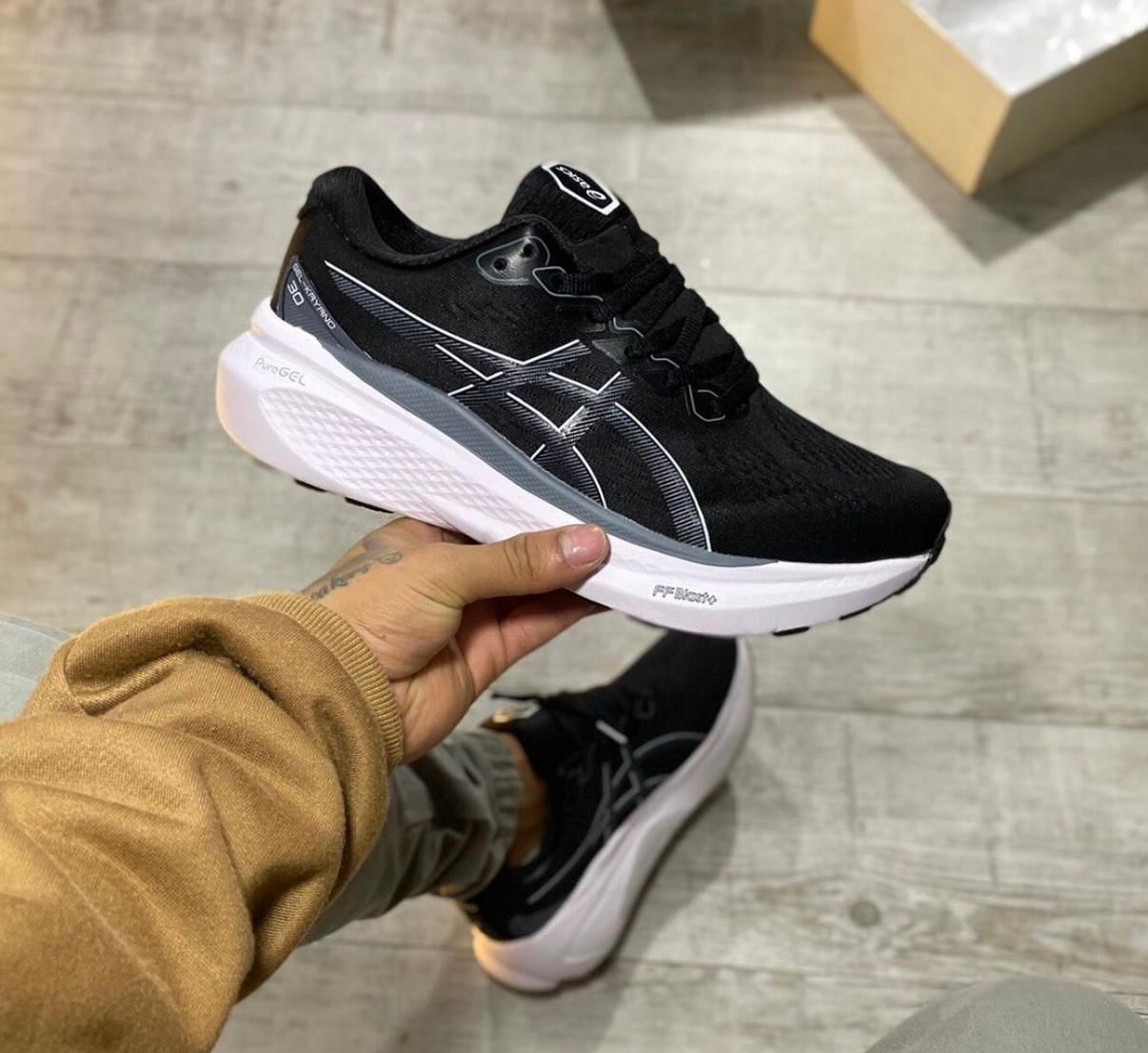 WhatsAppImage2025-05-14at16.07.54 | UAE SPORT STORE Black athletic running shoe with white sole and ASICS-style stripes, being held by a person indoors over a light wooden floor.