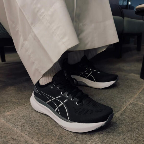 Person wearing black athletic sneakers with white soles, laces, and stitching, indoors on a tiled floor with light-colored pants and gray socks.