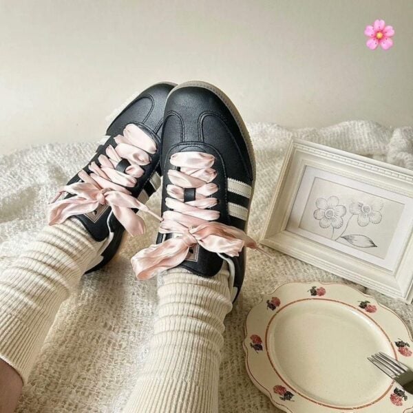 Black sneakers with pale pink ribbon laces resting on cream socks and a cozy textured surface.