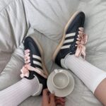 Navy sneakers with pink satin ribbon laces on a light gray sofa, a teacup and saucer nearby.