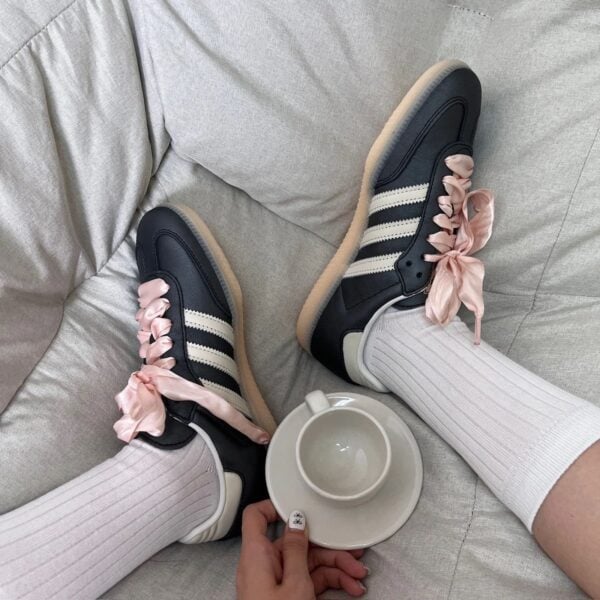 Navy sneakers with pink satin ribbon laces on a light gray sofa, a teacup and saucer nearby.