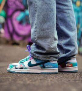 Close-up of colorful sneakers (turquoise, purple, white) with jeans, standing on pavement with a graffiti backdrop behind.