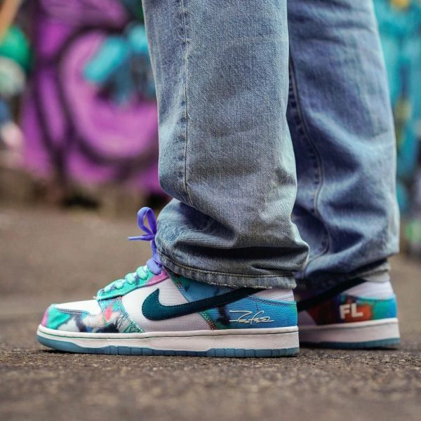 Close-up of colorful sneakers (turquoise, purple, white) with jeans, standing on pavement with a graffiti backdrop behind.
