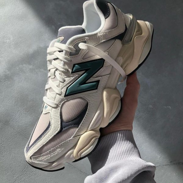 White athletic sneaker with teal New Balance 'N' logo held in a hand against a gray floor.
