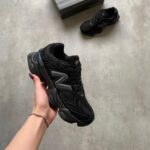 A black chunky sneaker being held in a hand, with another sneaker and a box on a gray floor in the background.