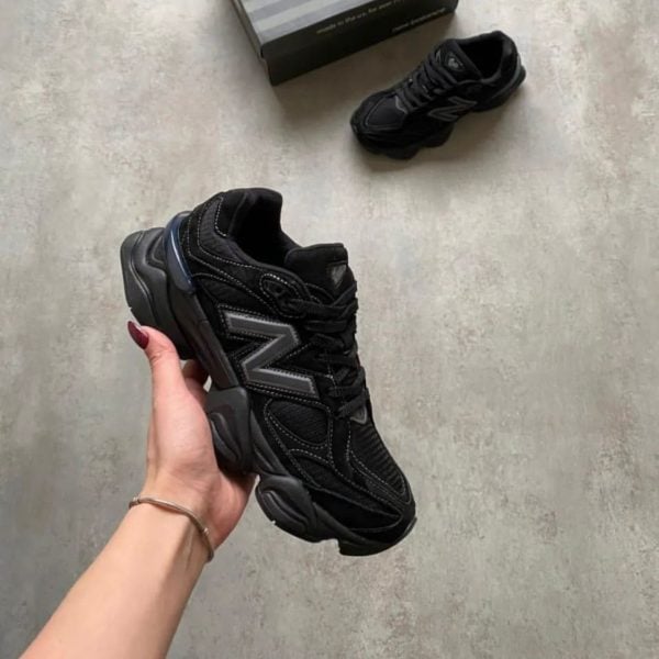 A black chunky sneaker being held in a hand, with another sneaker and a box on a gray floor in the background.