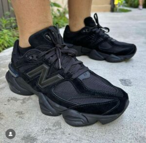 Black chunky athletic sneakers with thick soles on a person’s feet, standing on a concrete walkway outdoors.