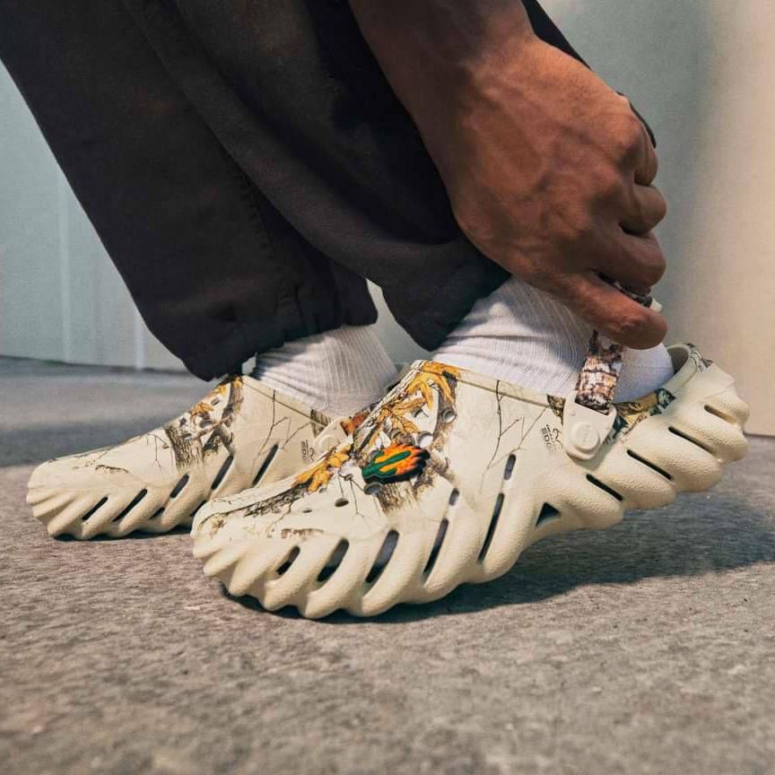WhatsApp_Image_2024-09-06_at_09.18.50 | UAE SPORT STORE Person adjusting off-white sneakers with cut-out, divided soles and tropical leaf/camouflage pattern, wearing white socks.