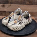 WhatsApp_Image_2024-09-06_at_09.18.55 | UAE SPORT STORE Pair of beige camouflage Crocs with ventilation holes, displayed on a black circular platform, tags attached to the shoe.