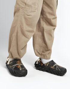 WhatsApp_Image_2024-09-06_at_11.31.56 | UAE SPORT STORE Lower body of a person in tan cargo pants wearing damaged black protective shoe covers with torn edges and white laces on a white studio floor.