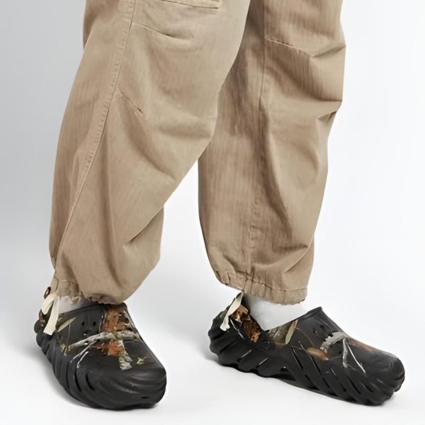 Lower body of a person in tan cargo pants wearing damaged black protective shoe covers with torn edges and white laces on a white studio floor.