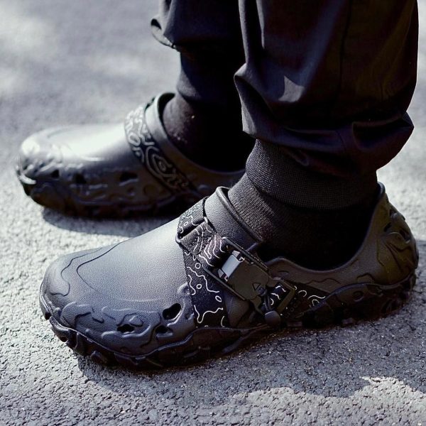 Close-up of black athletic shoes with patterned design and buckle on a person standing on asphalt surface.