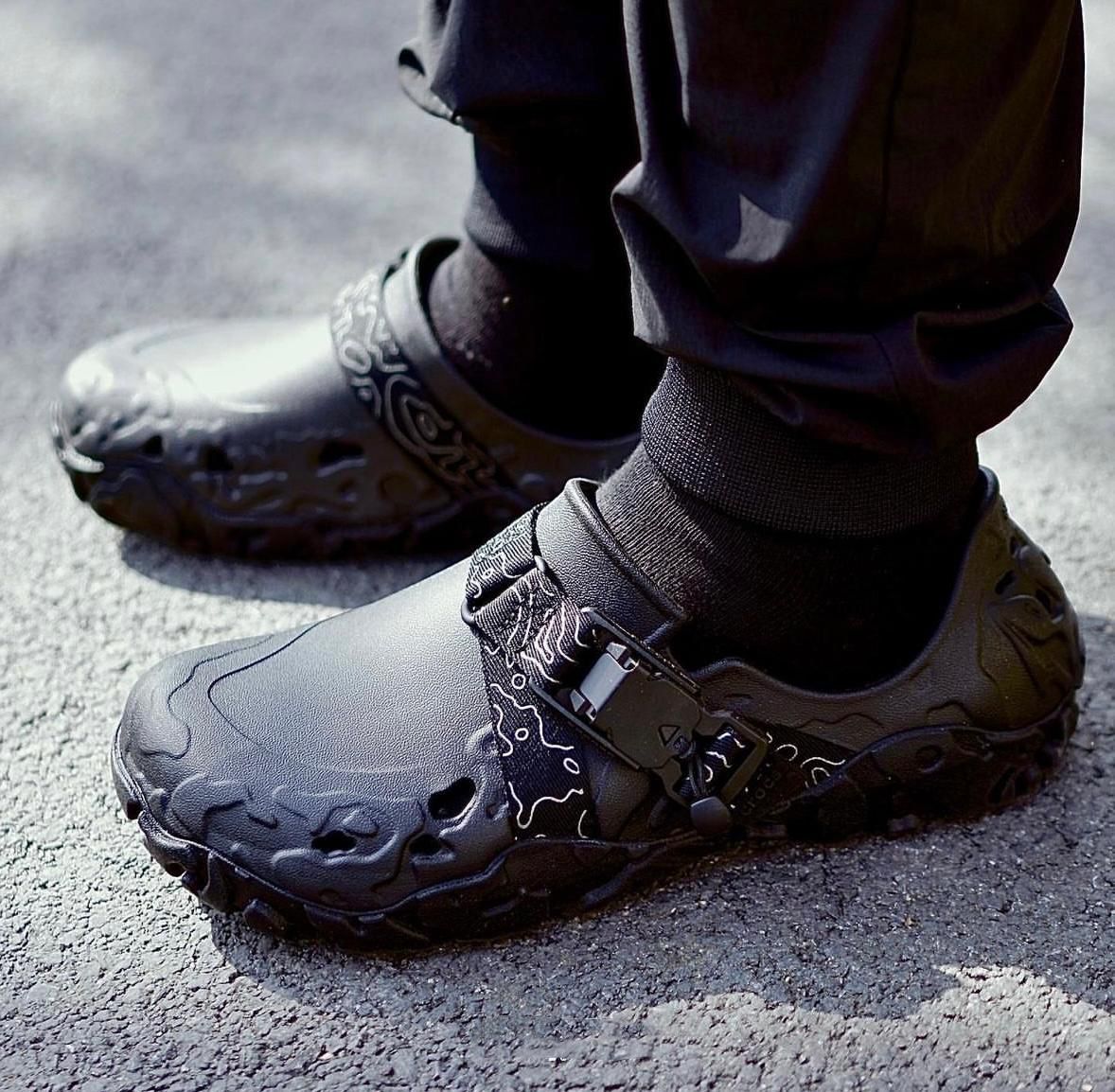 WhatsApp_Image_2024-09-07_at_09.58.10 | UAE SPORT STORE Close-up of black athletic shoes with patterned design and buckle on a person standing on asphalt surface.