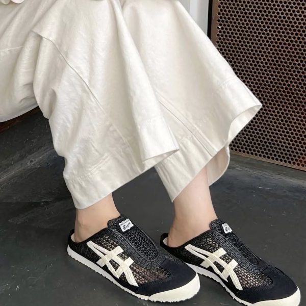 Person wearing black patterned sneakers with white stripes, standing on a dark floor, beige wide-leg pants draped around the ankles.