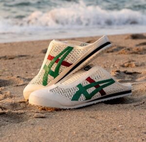 White mesh slip-on sneakers with green and red stripes resting on a sandy beach with waves in the background on the shoreline.