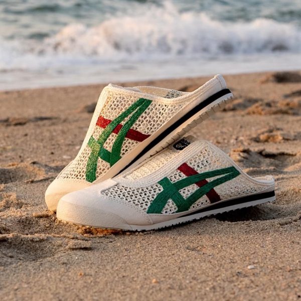White mesh slip-on sneakers with green and red stripes resting on a sandy beach with waves in the background on the shoreline.