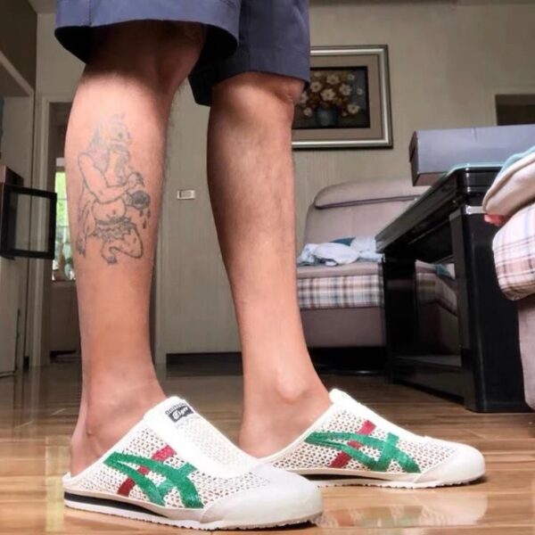 Person standing indoors wearing white mesh slip-on sneakers with green and red side stripes; tattooed left shin visible, living room with couch and bed in the background.