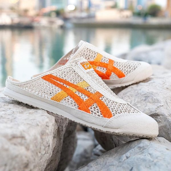 White mesh slip-on sneakers with orange stripes resting on gray rocks by a waterfront.