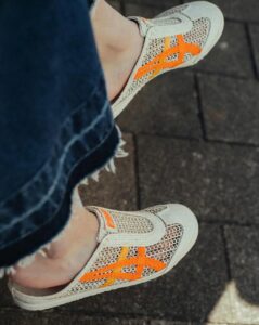WhatsApp_Image_2024-10-04_at_12.40.25 | UAE SPORT STORE Beige mesh sneakers with bright orange laces on a paved sidewalk, visible denim jeans cuffed at the ankle.