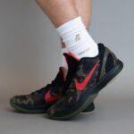 Foot wearing a black camouflage Nike basketball shoe with a red swoosh and a white sock visible above it, set against a gray background.