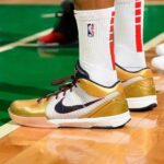 Gold and white Nike basketball shoes on a wooden court with white socks featuring NBA branding, black swoosh logo visible.