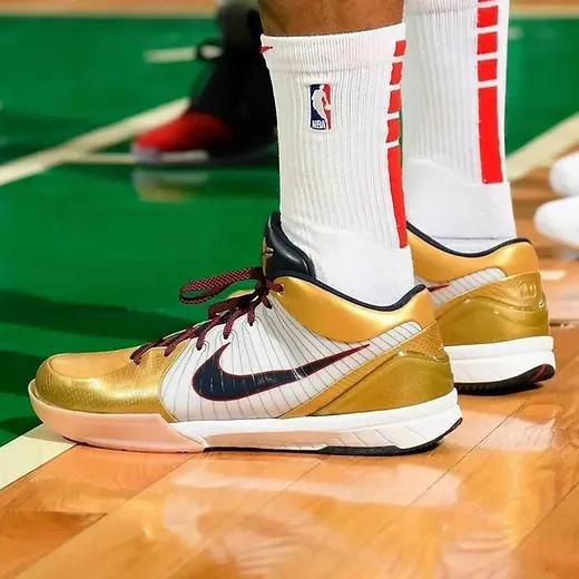 Gold and white Nike basketball shoes on a wooden court with white socks featuring NBA branding, black swoosh logo visible.