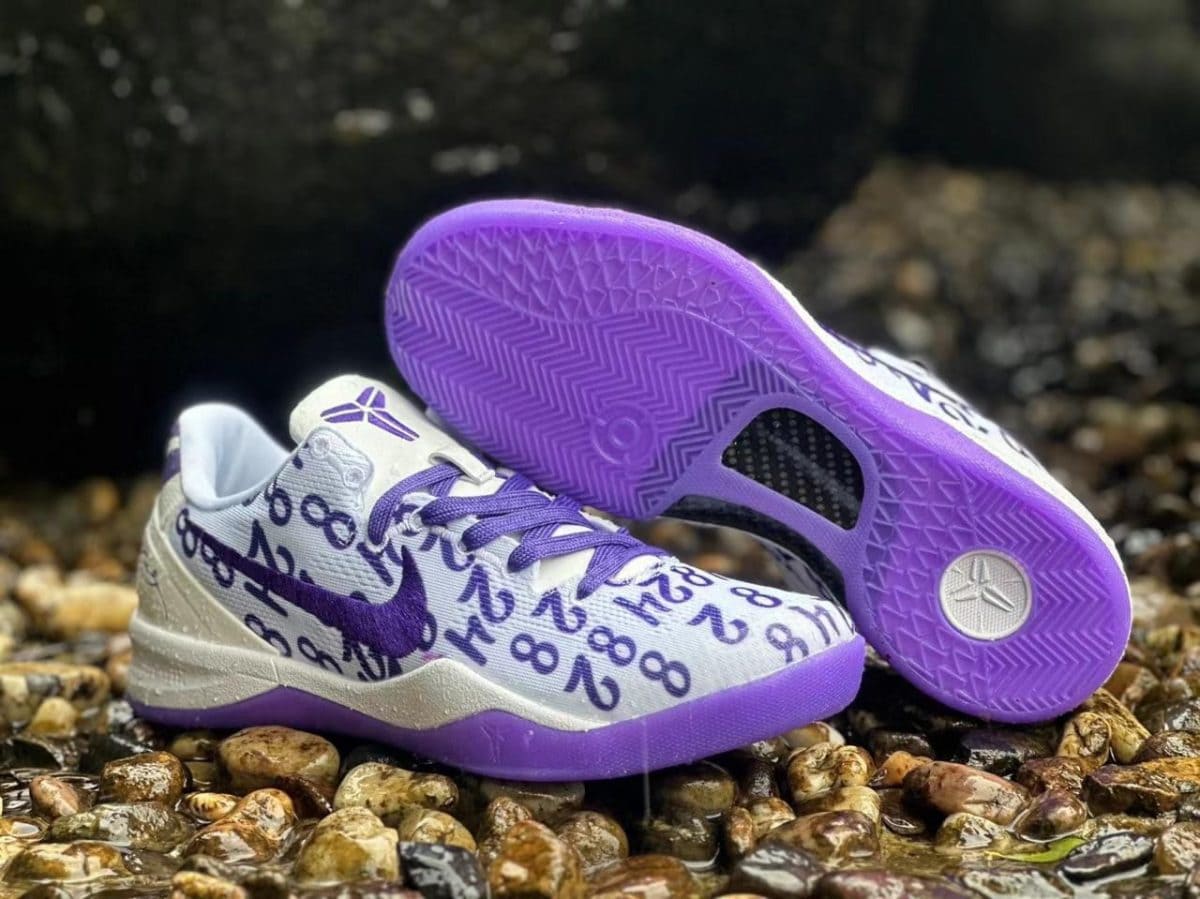 White and purple athletic shoes with a numeric pattern, resting on wet pebbles by a dark background.