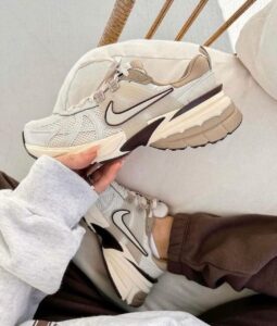Cream and white Nike running shoes with black swoosh being held up, multiple pairs visible on a light couch backdrop.