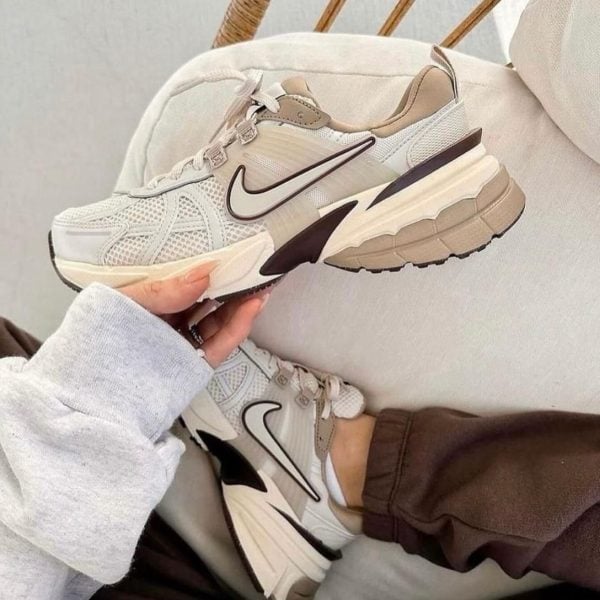 Cream and white Nike running shoes with black swoosh being held up, multiple pairs visible on a light couch backdrop.