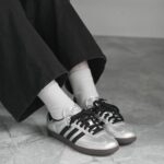 Person wearing metallic silver sneakers with white socks and black pants standing on a tiled floor (close-up of feet).
