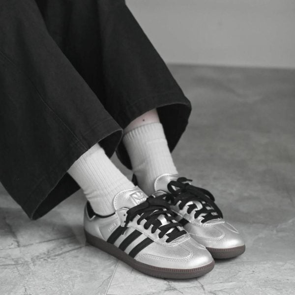 Person wearing metallic silver sneakers with white socks and black pants standing on a tiled floor (close-up of feet).