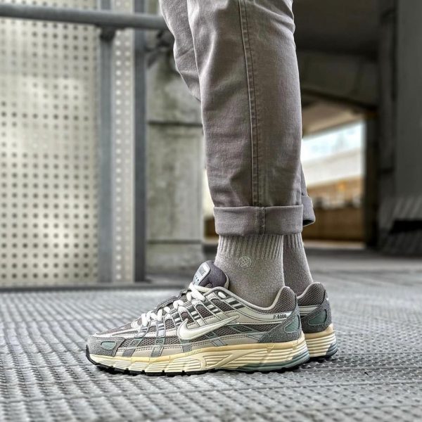 Side view of a gray Nike sneaker on a metal grate, with rolled gray jeans and matching socks in an urban setting (P-6000 visible).