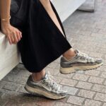 Person in a black dress sits with legs extended, wearing gray running sneakers on a brick sidewalk.