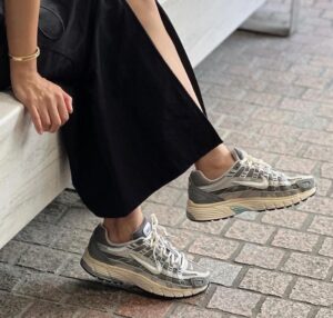 Person in a black dress sits with legs extended, wearing gray running sneakers on a brick sidewalk.