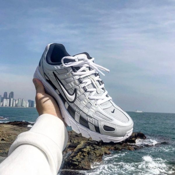 White and gray Nike running shoe held up by a person in a white sleeve, with the ocean, rocks, and distant city skyline in the background.