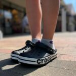 WhatsApp_Image_2024-10-18_at_11.29.24 | UAE SPORT STORE Person wearing black Crocs with a white sole and gray Champion socks standing on a brick-paved sidewalk outdoors.